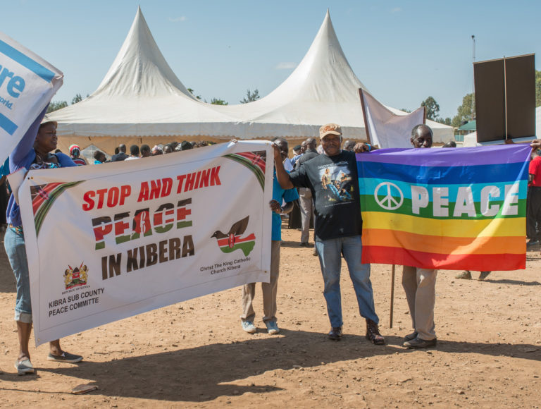 Peace March in Kibera with signs saying "Stop and Think Peace in Kibera" and "Peace" on a rainbow background with a peace sign.