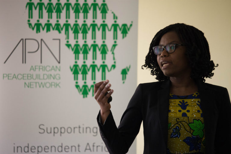 Dr. Peace Medie giving a lecture with an APN banner in the background