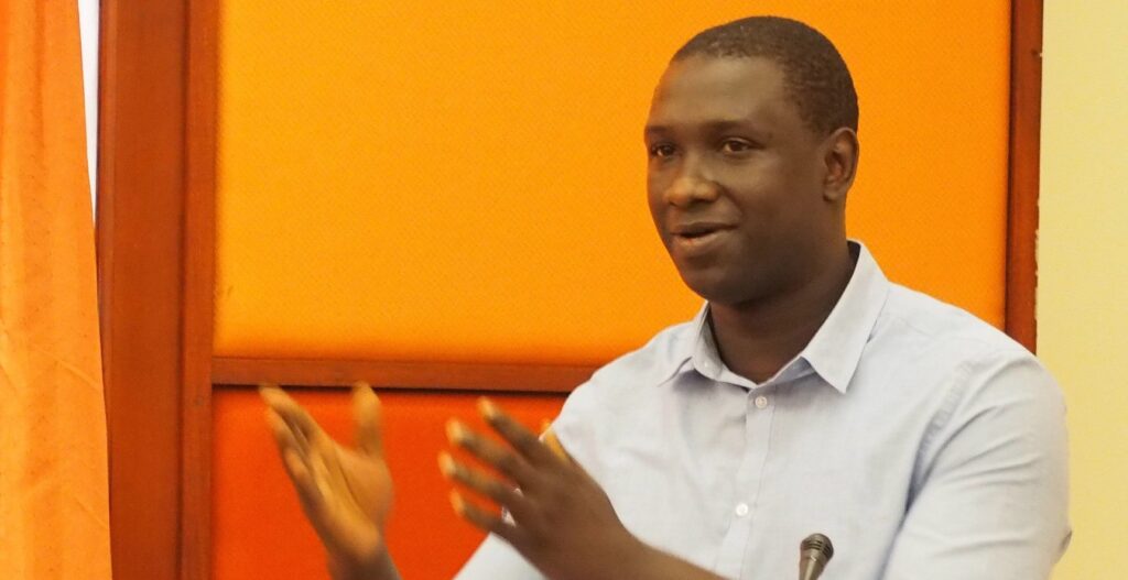 Dr. Ibrahim Bangura giving a presentation. Both hands are extended and there is an orange wall in the background.