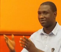 Dr. Ibrahim Bangura giving a presentation. Both hands are extended and there is an orange wall in the background.